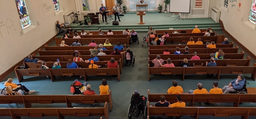 Looking down on a row of church pews with a lot of people sitting in them or next to them in wheelchairs. 