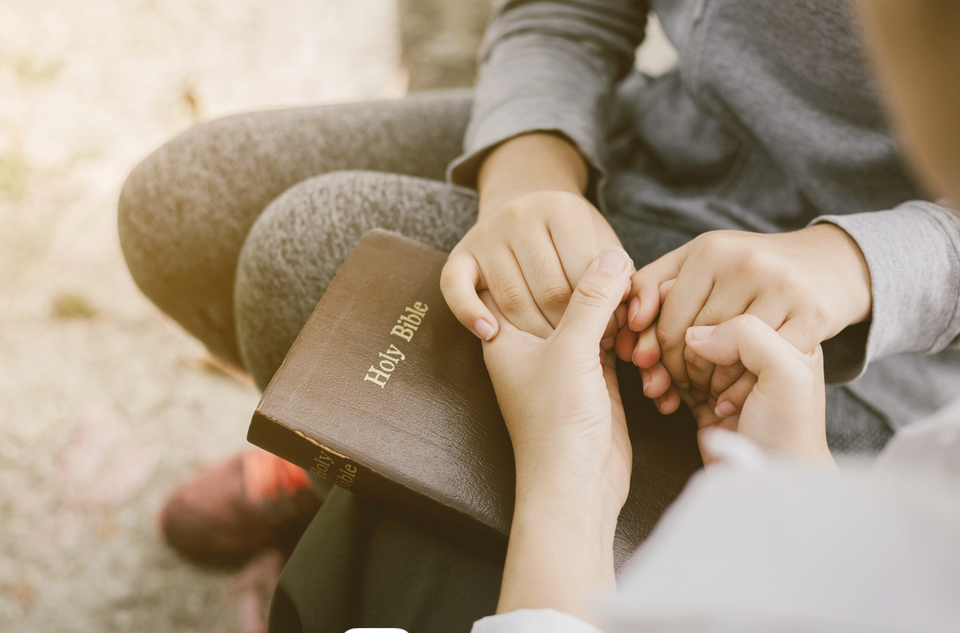 Two people holding hands praying with bible