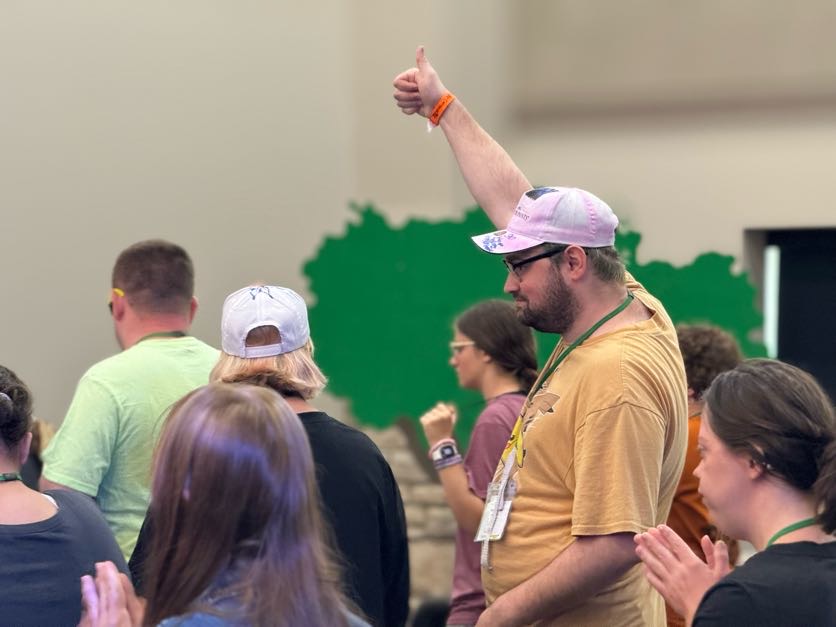 Man with thumb up held in the air over his head in a room full of people