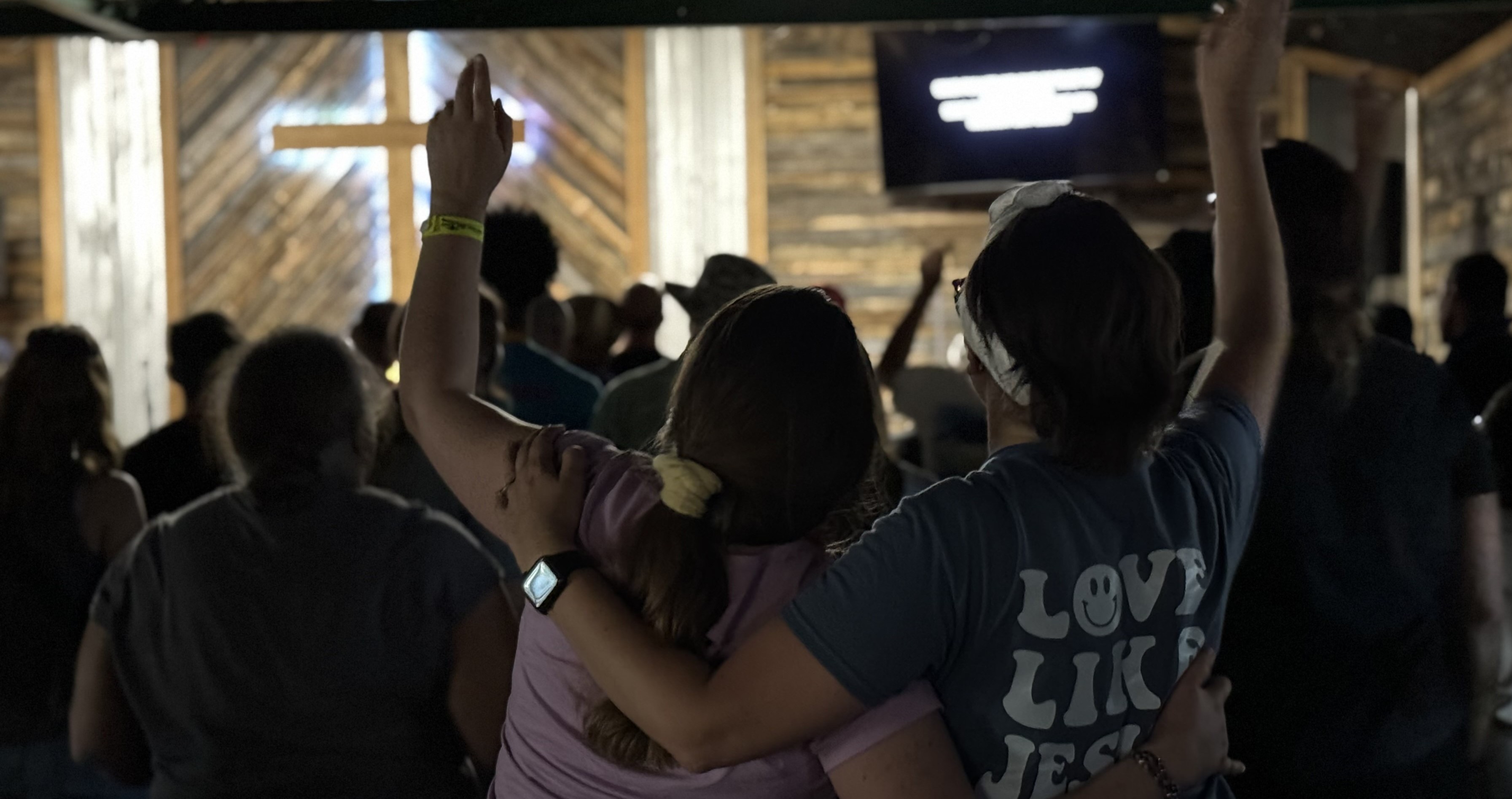 Two people facing away praising God with a lit cross at stage in front of a crowded room