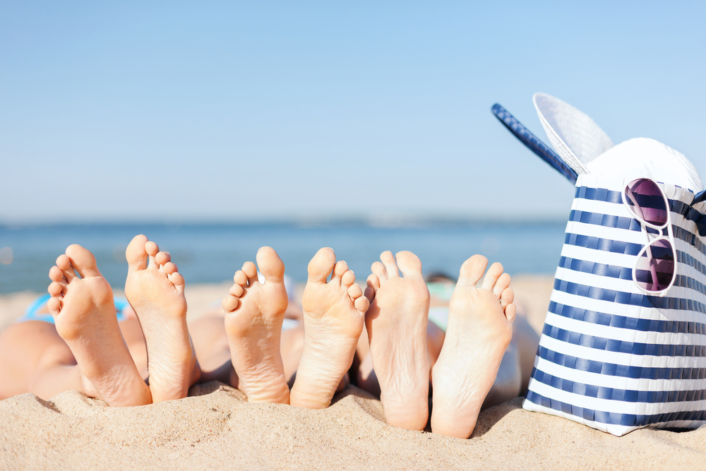 3 pairs of feet on a beach next to a bag