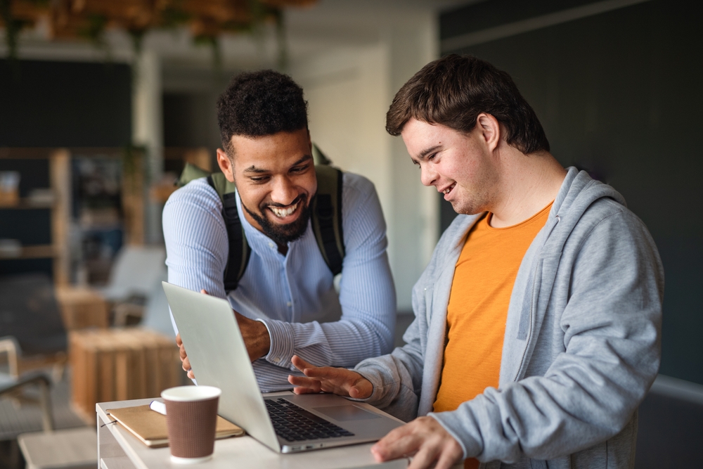 Two guys one with a disability looking at a laptop and smiling