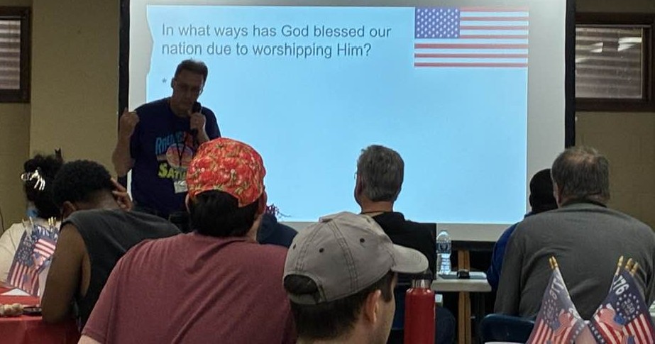 A presenter speaking with a projection behind him reading In what ways has God blessed our nation due to worshipping Him?