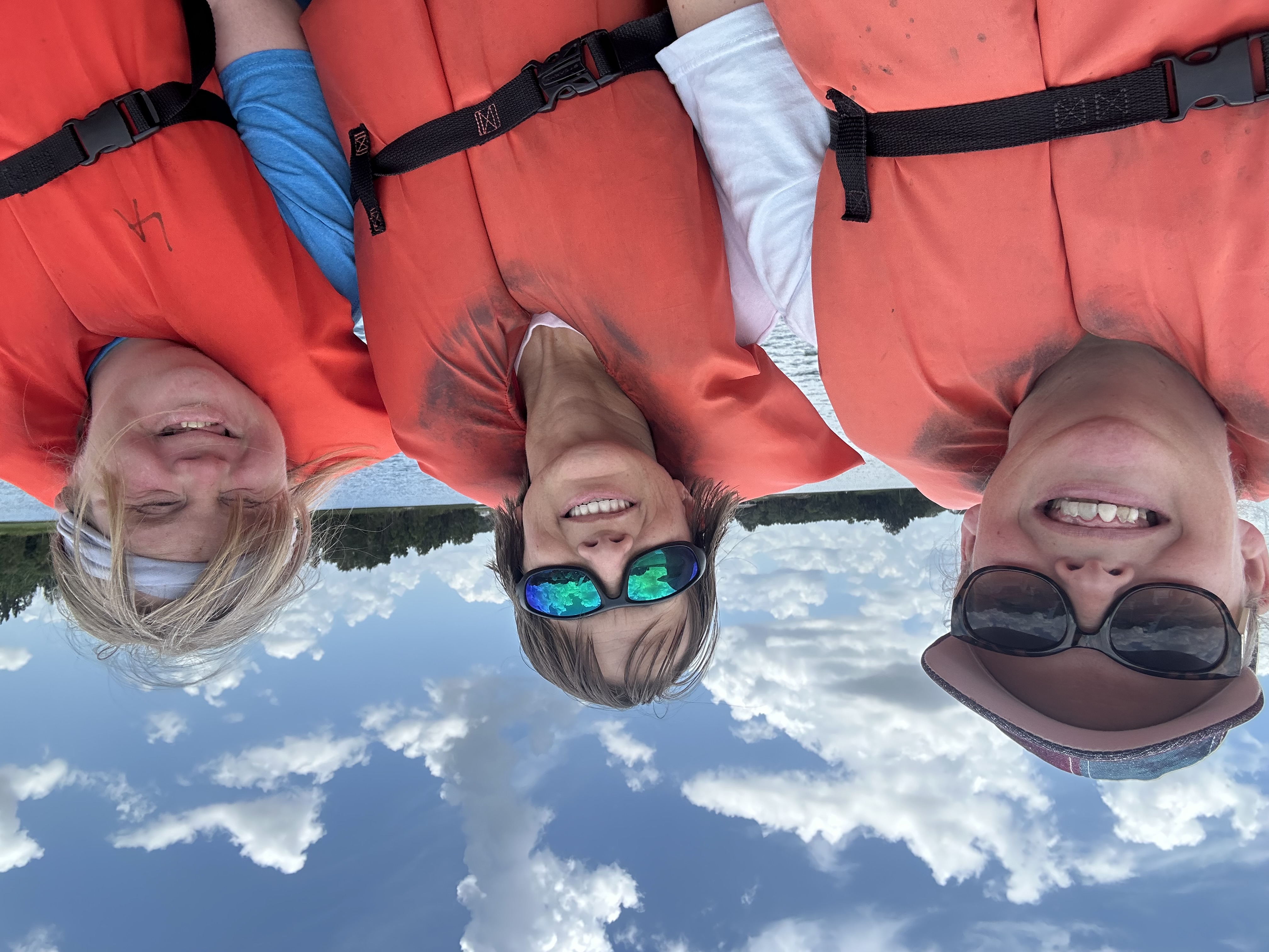 three people wearing life jackets smiling while on a boat on the water under a sunny blue sky
