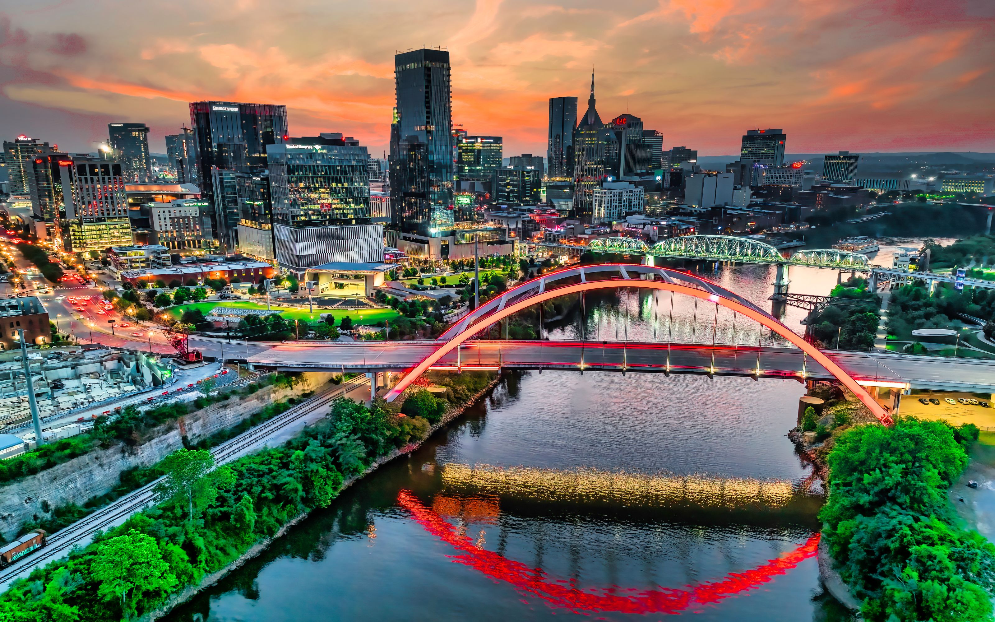 A skyline of Nashville, TN at sunset. The foreground includes a bridge over a river, and the city skyline is in the background with an orange and grey sky overhead.