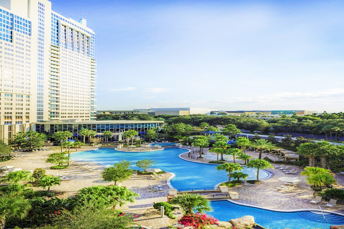 On the left, a large hotel rises toward the sky. A blue and white sky with light clouds takes most of the image. The bottom half is a bright blue pool with palm trees.