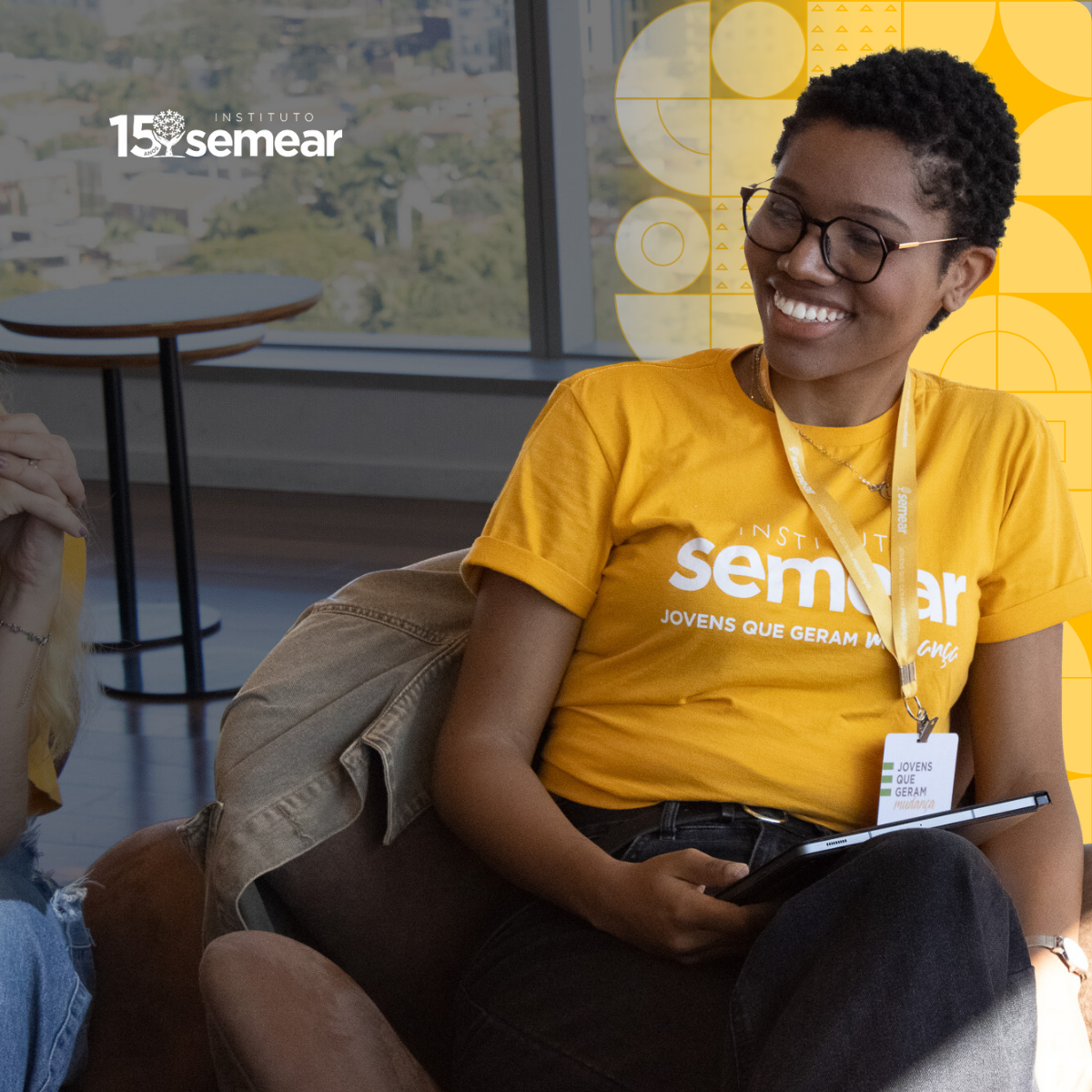 Uma jovem sorridente com camiseta amarela faz o sinal de paz; ela usa um crachá e está em frente a um fundo amarelo com ícones de livro e capelo (chapéu de formatura).