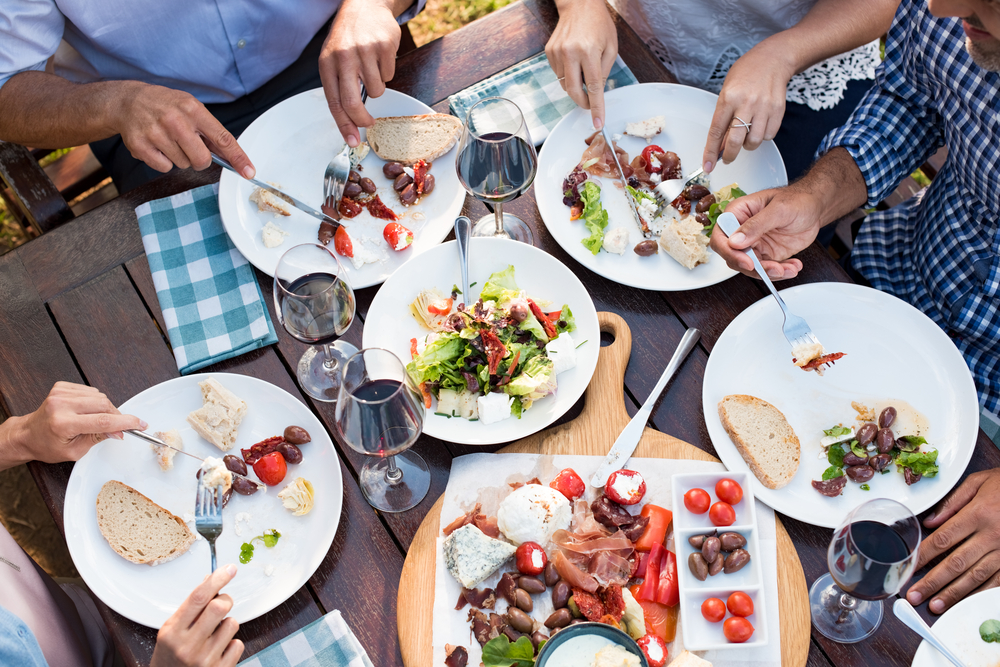 Friends enjoying eating lunch together outdoor. Top view of mature friends hands having lunch with wine and salad. High angle