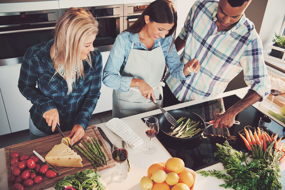 High angle view of trio cooking a meal with tomatoes, cheese, asparagus, fruit and other ingredients on kitchen counter and stove