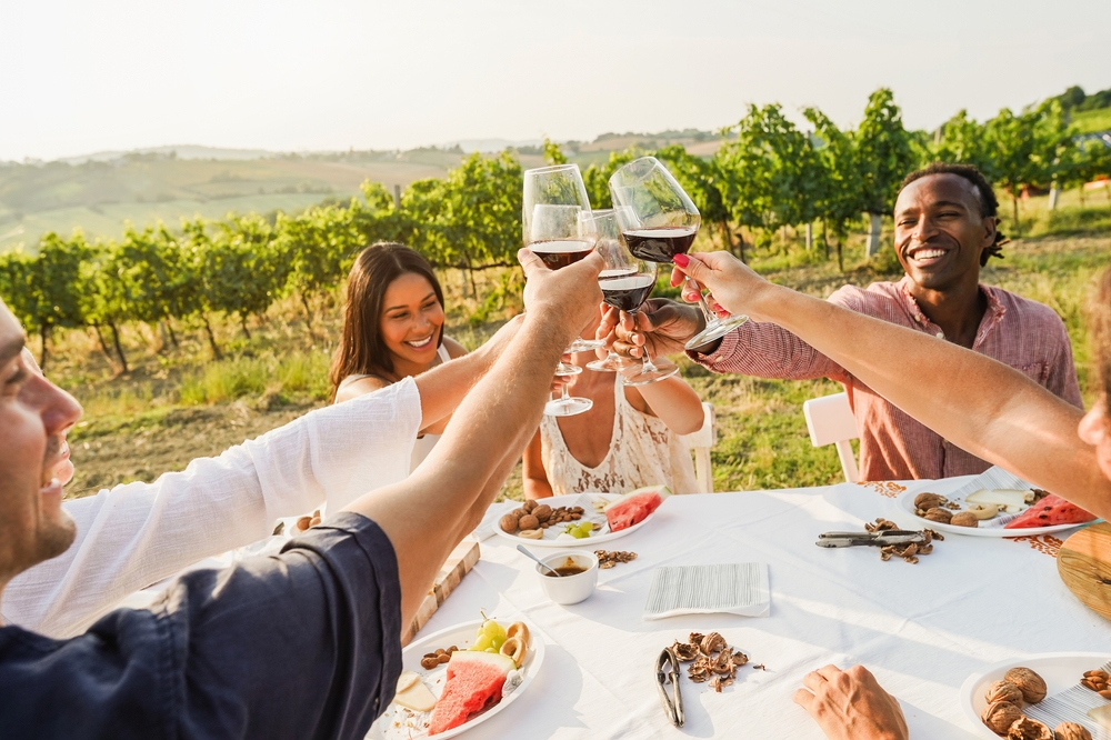 Happy adult friends toasting wine glasses at a table in a vineyard in Tuscany