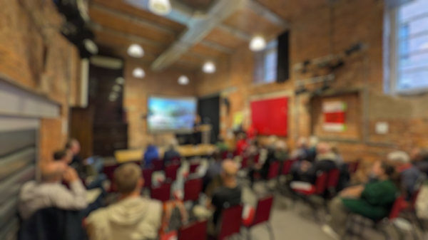 A blurred photo from the Carbon Co-op AGM of a room full of chairs and people facing towards a stage