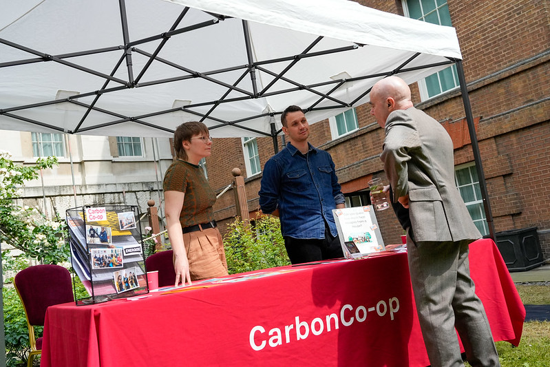 Two Carbon Co-op staff members (Ellie Radcliffe & Pete Fletcher) standing behind a table with a red cloth saying “Carbon Co-op“ on it, speaking to a man in a suit in the garden of Number 10 Downing Street
