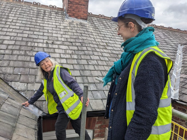 A photo of two women in high vis jackets and helmets standing on scaffolding at roof level