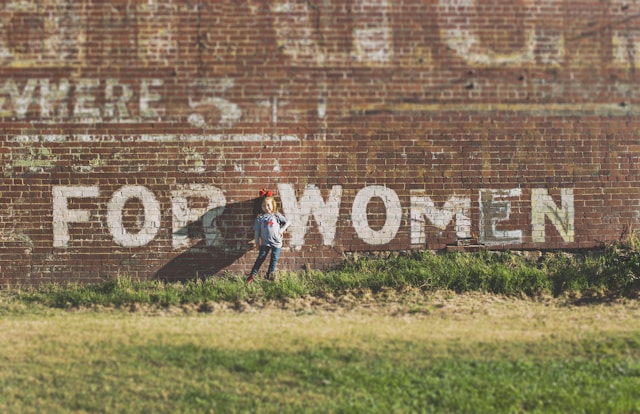 A photograph of a young girl standing in front of a brick wall with the words 'FOR WOMEN' painted on in large white letters.