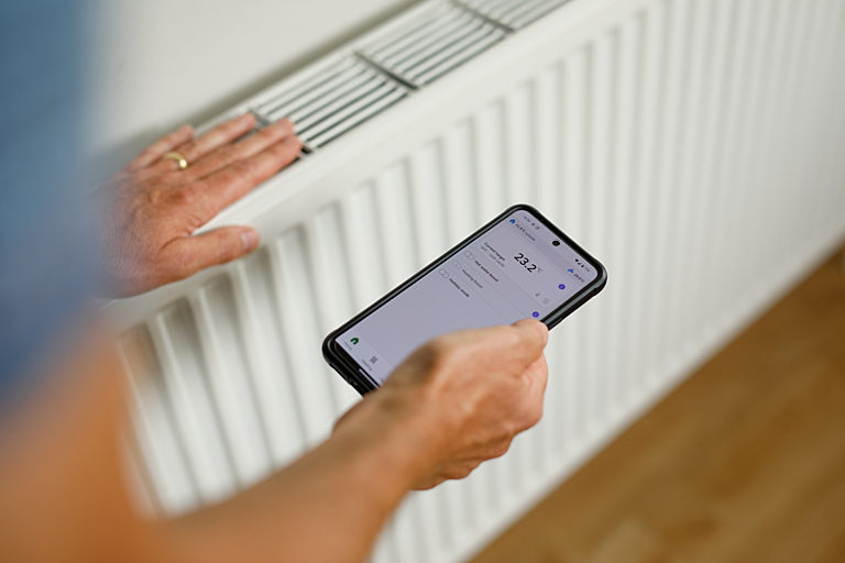 Dan Curran uses his phone whilst connected to a heat pump system at home in Surbiton, Greater London, UK, 18th August 2025. ..<br>..<br>..Dan Curran, a supply chain and logistics consultant, lives in a 1930’s, solid brick detached family home in Surbiton, South West London. Interested in sustainable living and improving his environmental footprint, Dan installed a heat pump, a Samsung 12kW R290 Monobloc, in 2024, along with solar panels and a battery. ..<br>..<br>..Since its installation, the heat pump has “created a more consistent and nice temperature” than the familiar hot and cold cycle of gas heating. How has living with a heat pump affected his life? “In a sense it hasn’t because it just works” he says, “it shouldn’t be seen as a life-changing decision” before adding “on the other hand I'm a bit obsessed with it!”...<br>..<br>..These photographs are part of a new resource, produced by Climate Visuals and Nesta, of compelling, engaging and accessible images of heat pumps across the UK. All too often, photographs of heat pumps show the object but don’t tell the story. They depict a box at the side of a building, often with awkwardly posed models standing beside it. They don’t show the real world potential of heat pumps that audiences can relate to and see themselves as a part of, or the diversity of installers and users. These new images highlight the real lives that heat pumps are a part of now, helping to normalise the technology and demonstrate the benefits being felt by the households using them across different houses and lifestyles around the country.