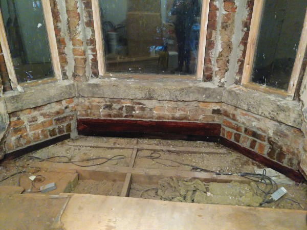 A photo of a bay window with plaster removed to expose brickwork and floorboards lifted
