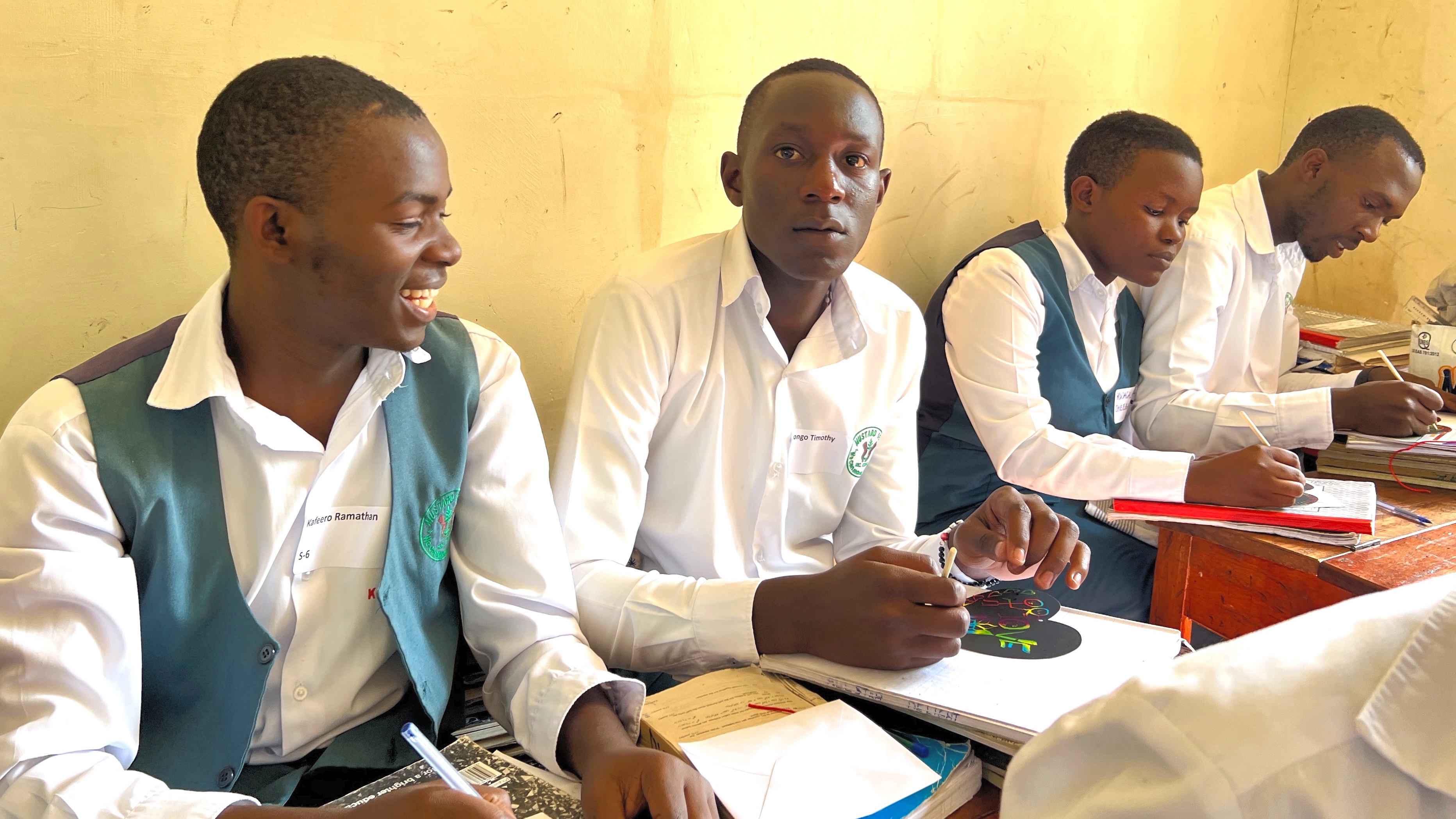 Four young adult Ugandan students crowded into two small desks.