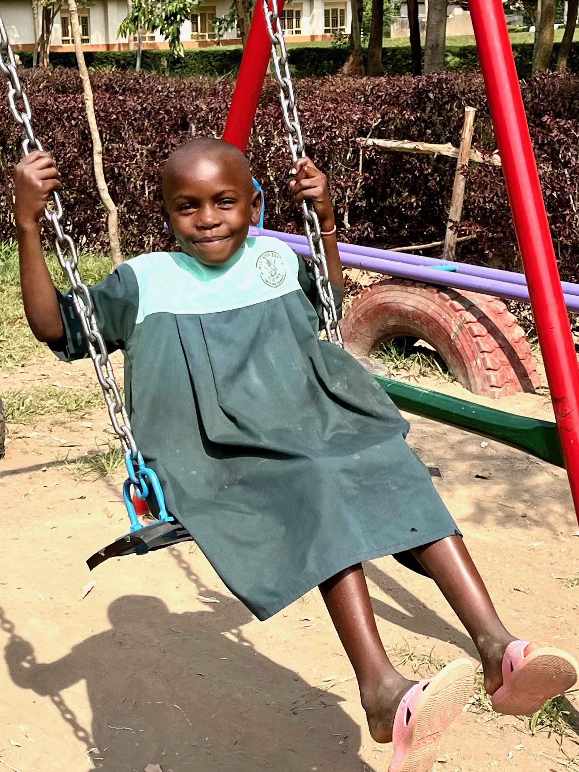 a young child swinging on a playground swing set, smiling brightly at the camera. The child is wearing a green school uniform and pink sandals. The swing set has red metal poles, and there are various other playground items visible in the background