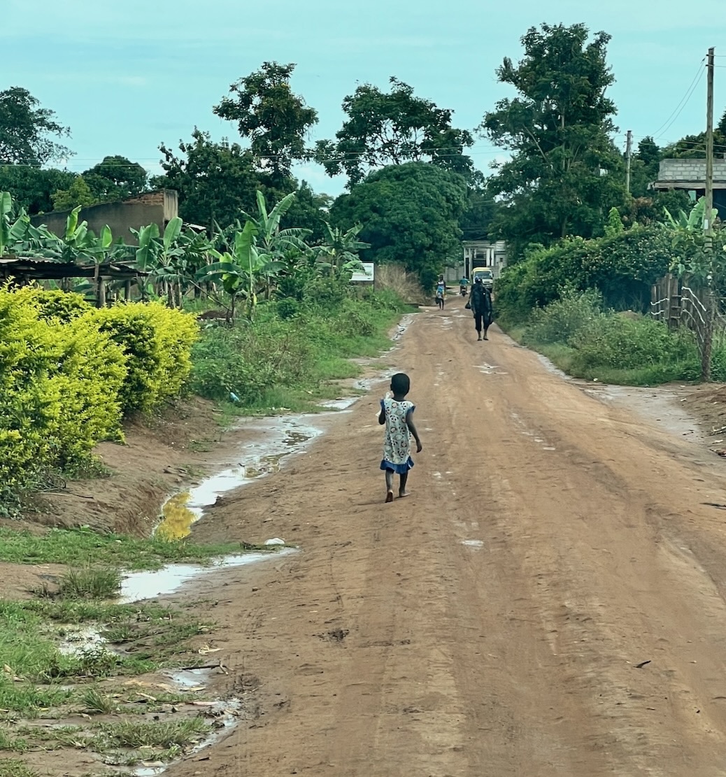 a little bafe-footed girl is walking up a narrow, muddy, dirt road. Lots of green plants on both sides of the road, and a few simple buildigs. In the distance, there is a man walking towards her. And there's a motorcycle and a van. Otherwise, it looks quite isolated.
