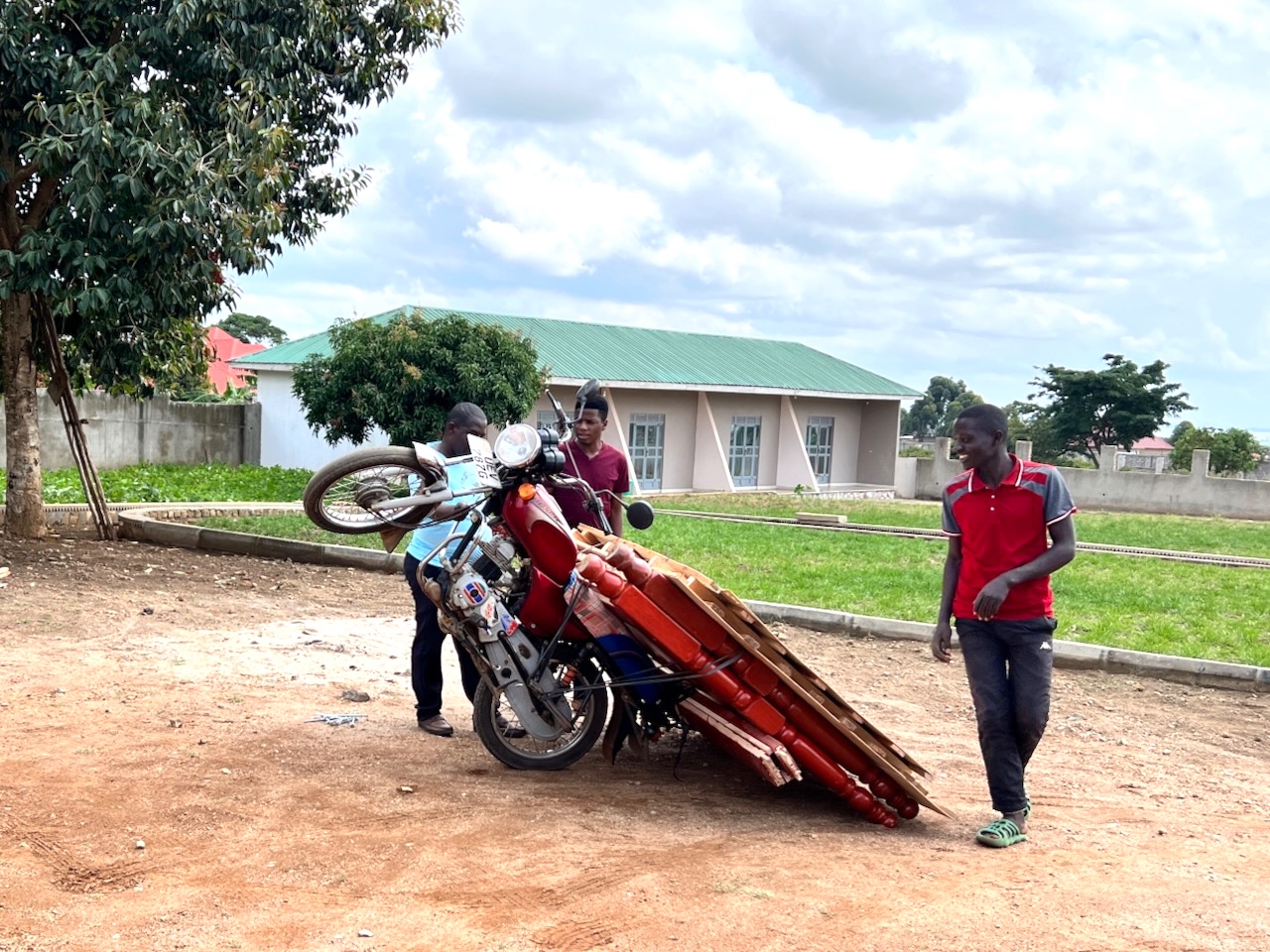 Three men with a motorbike tipped back on one wheel and big plywood sheets and rope resting on the bike and on the ground. 