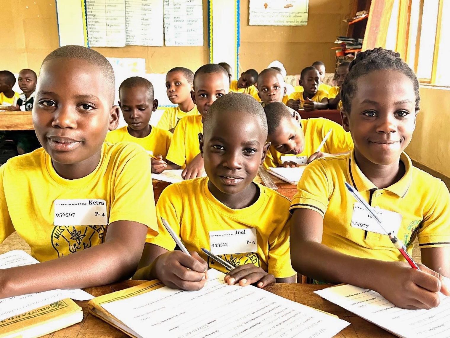 Classroom filled with bright, enthusiastic-looking Ugandan students in sunshine yellow shirts,  writing on a large piece of paper.