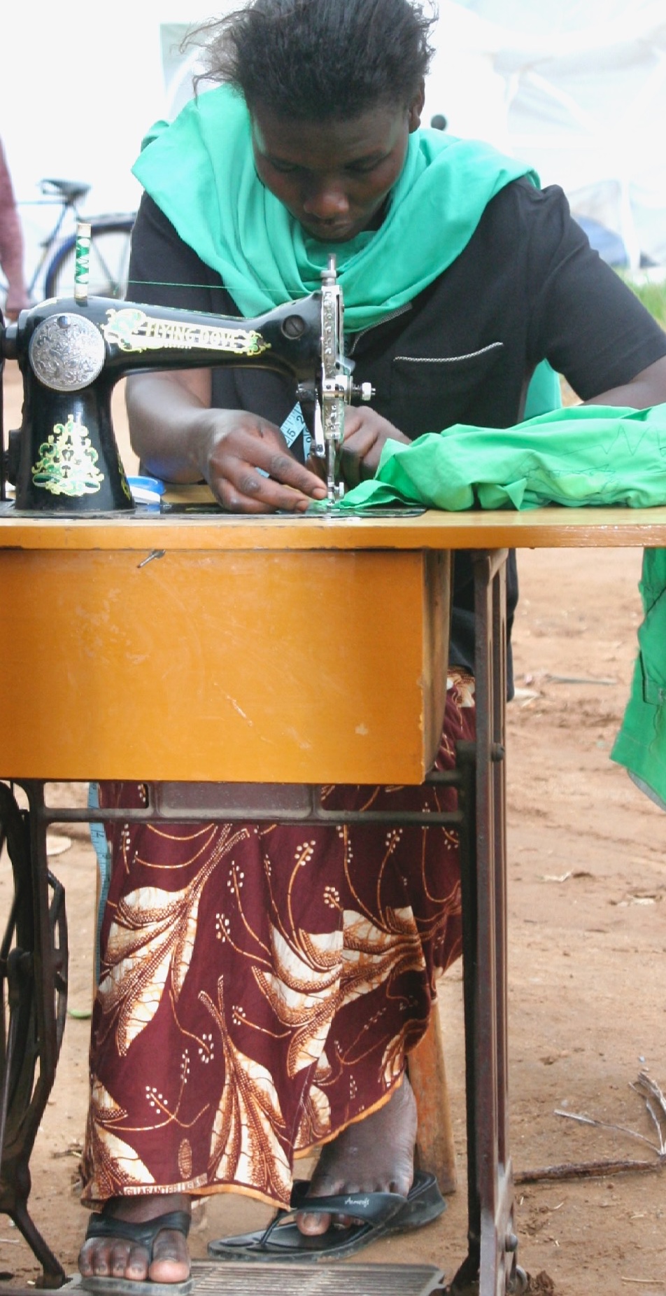 A Ugandan woman sitting at a treadle-style sewing machine along the side of the road while mending green fabric clothing