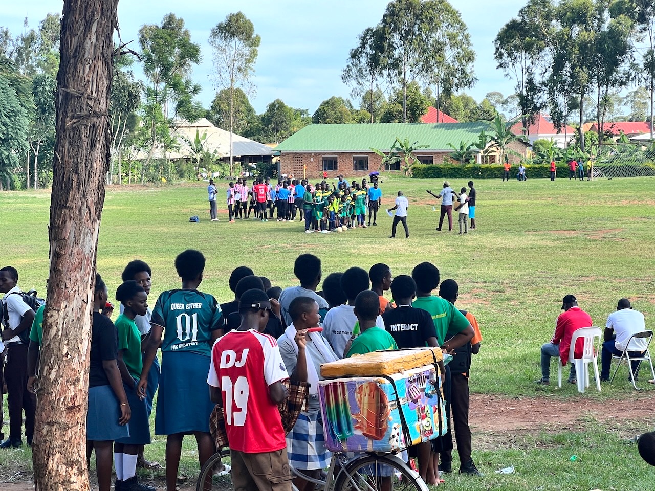 the foreground of the photo is a small crowd of students, a bicycle aith a cooler strapped on it (and painted with pictures of popsicles. In the middle of the photo are two foodball teams, about to begin the game. In the background are trees and a brick building with a green roof. 