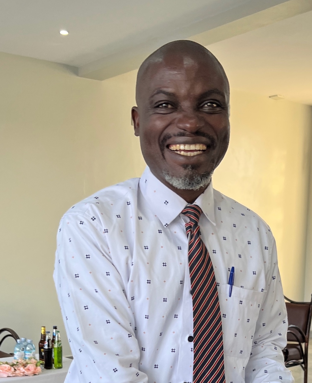 An older Ugandan man in a white lab coat next to a school building and a tree