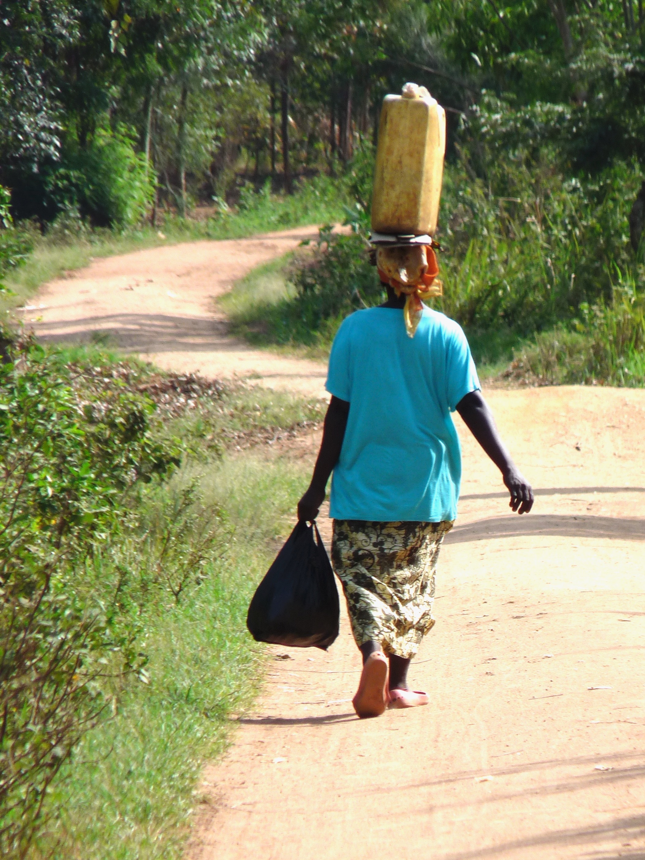 A Ugandan woman walking down a winding road with a 5-gal. jerry can of water on her head.