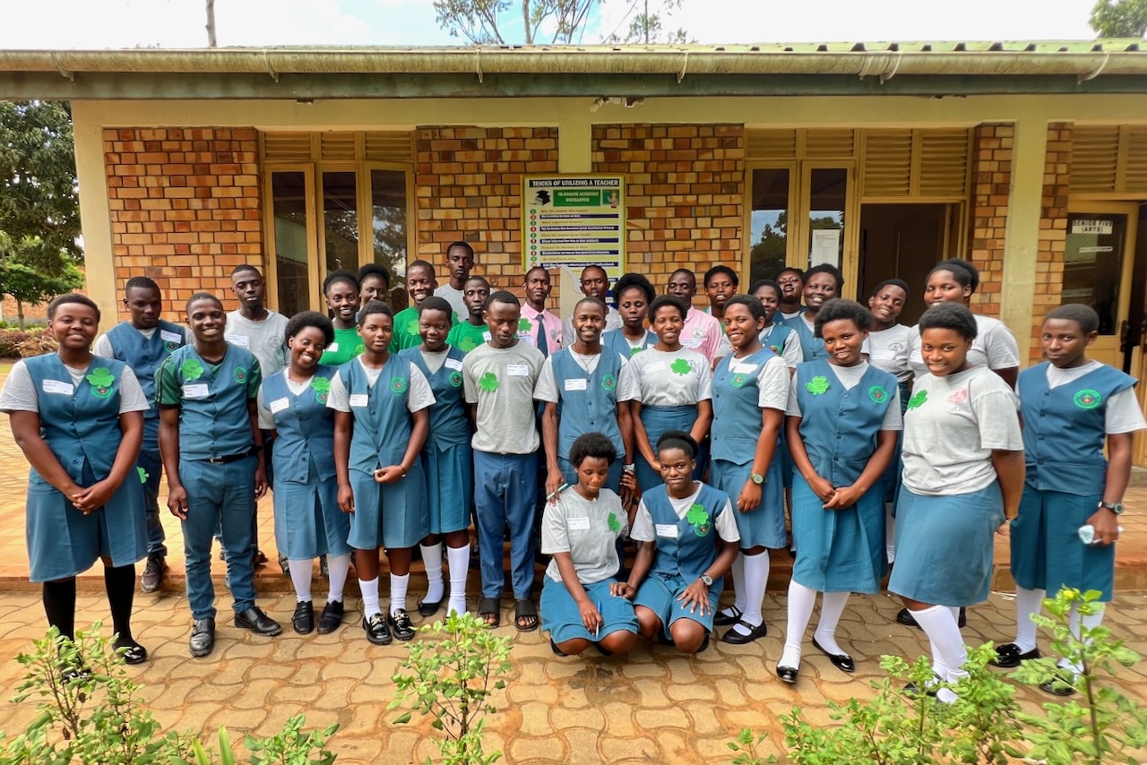 A group of Ugandan high school students, dressed in teal green uniforms in front of a classroom building, looking happy and confident.