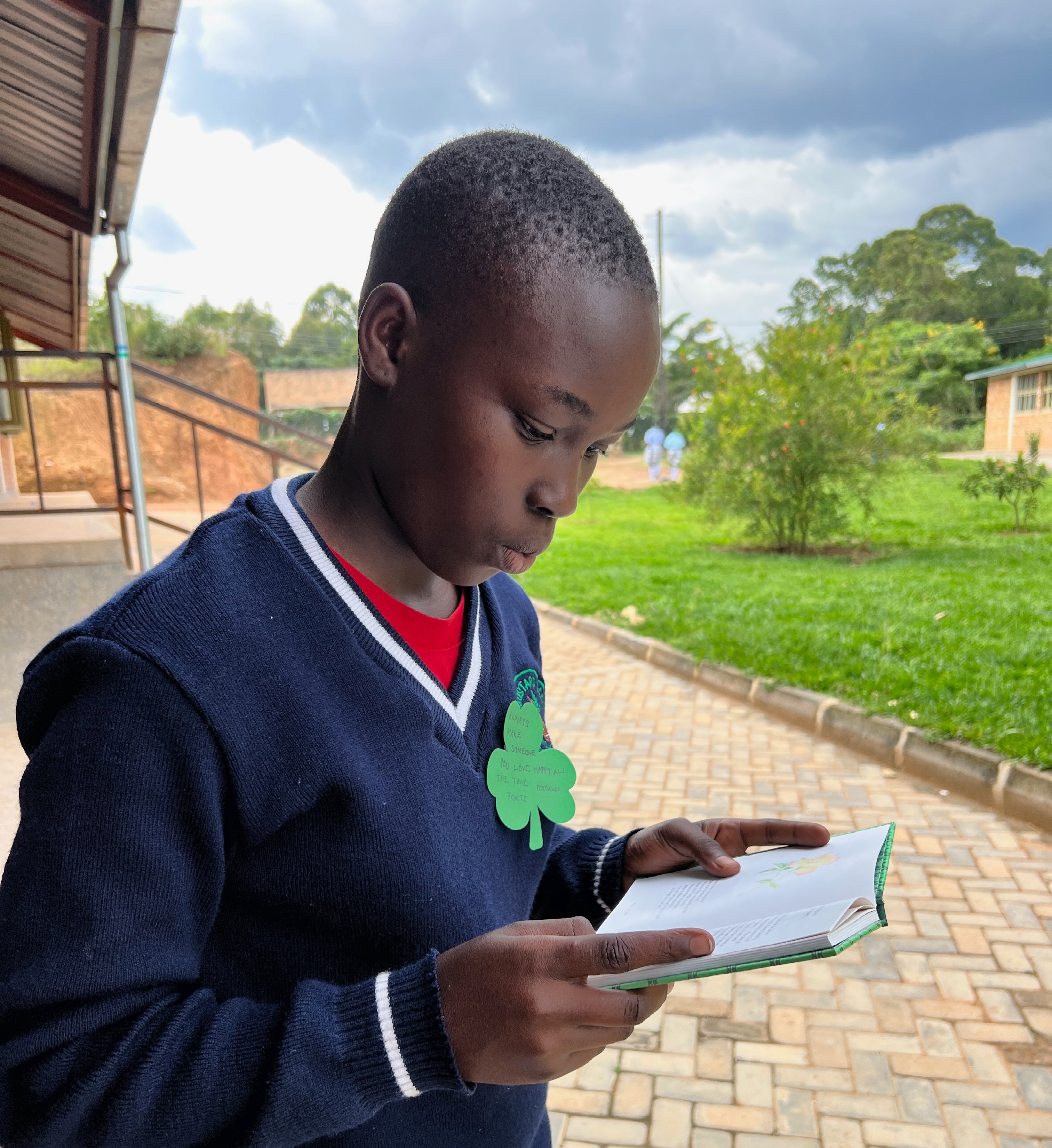 A sixteen year old Ugandan student reading a book with the school campus in the background
