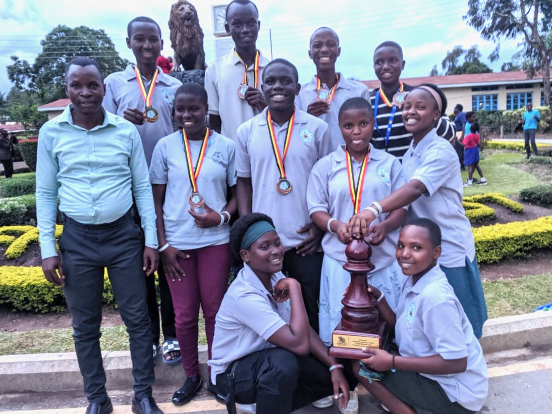 Ten Ugandan high school stuents with one teacher. They are holding a big trophy shaped like a chess piece, and many are wearing medals. Behind them are some school buildings and an area with a statue of  the school's mascot, a lion.  