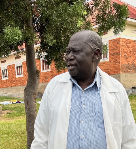 An older Ugandan man in a white lab coat next to a school building and a tree