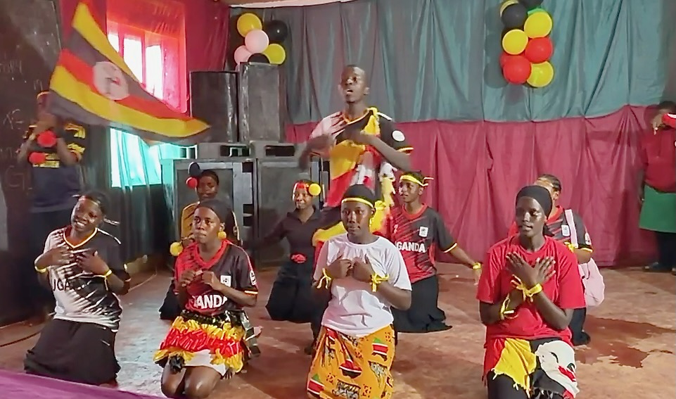 A colorful background with the colors of the Ugandan flag- yellow, red, and black. Eight teen-age girls kneelig, each dressed in a variety of red, yelow and black costumes. Two youg men- one waving the Ugandan flag, and one, singing and directing the dancers. 