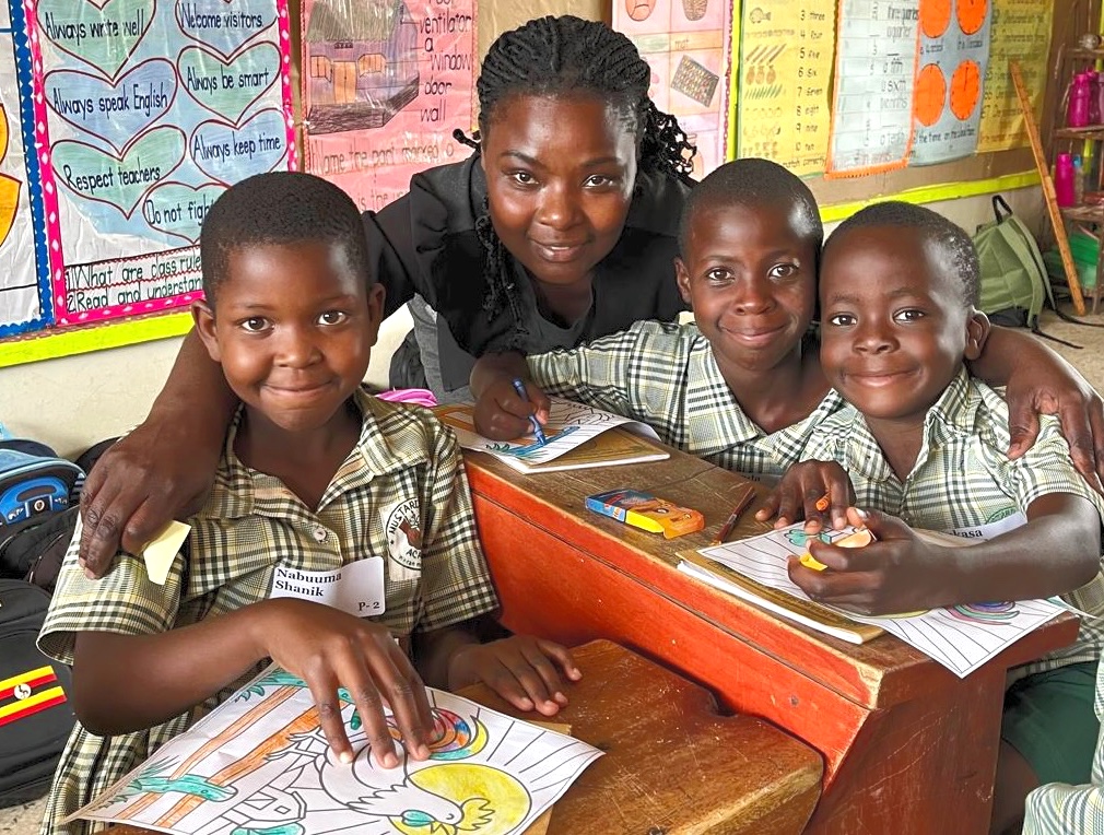 three Primary-2 (second-graders) and their teacher, who has her arms around them. They are all smiling and looking very comfortable and happy. On the desks are coloring pages and crayons. The children have colored a farmyard scene with a very colorful rooster with the sun rising behind him. In the background are posters. One of them has class rules: “Always write well. Welcome visitors. Always speak English. Respect teachers. Always be smart. Always keep time. Do not fight.“