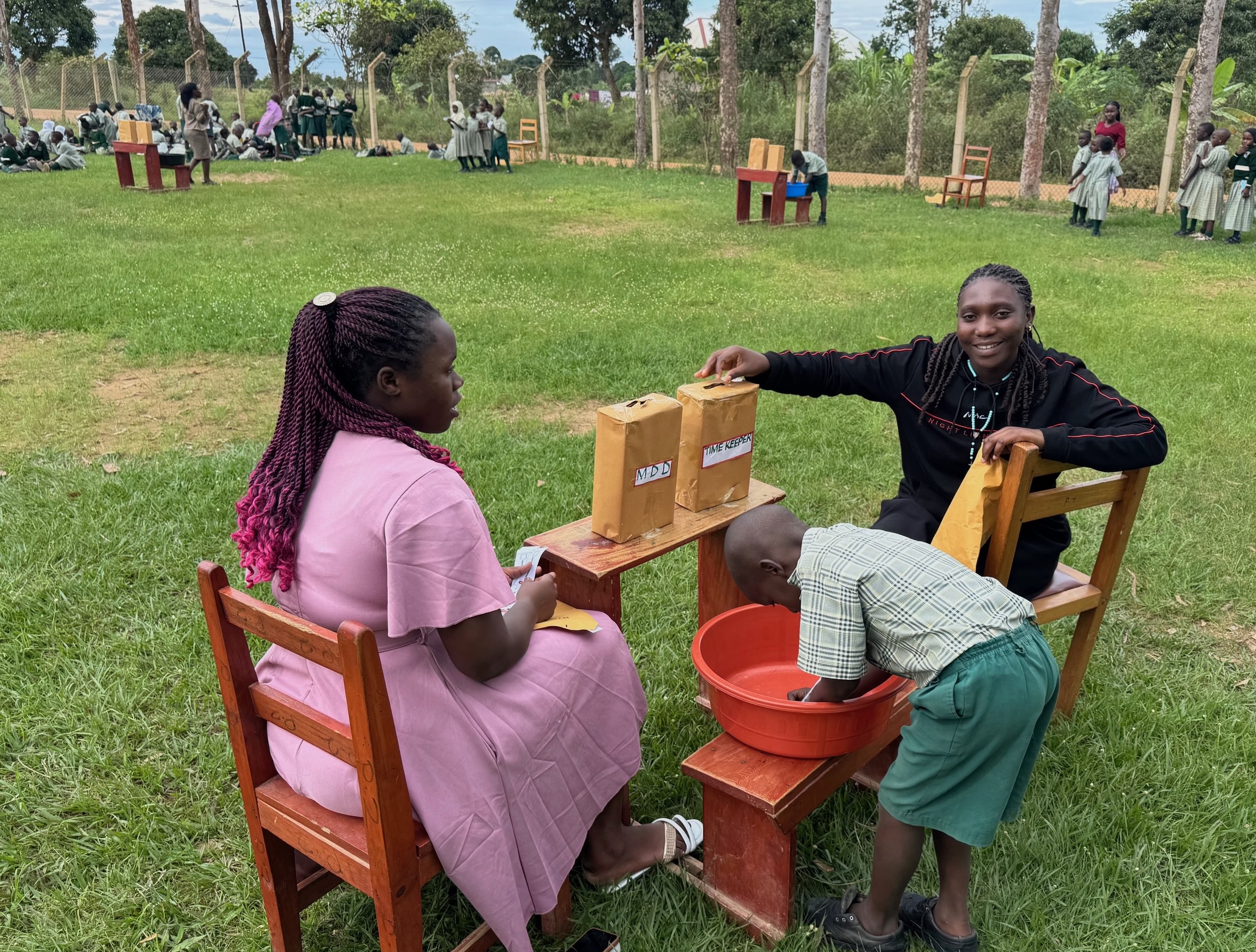 the foreground of the photo is a little boy dressed in a green school uniform, bending to write inside an orange basin. There is a school desk with two ballot boxes, one for “Timekeeper“ and one for “MDD.“ On either side of the desk is a teacher. In the background, you can see two more setups with ballot boxes and queues of students waiting to cast their votes.