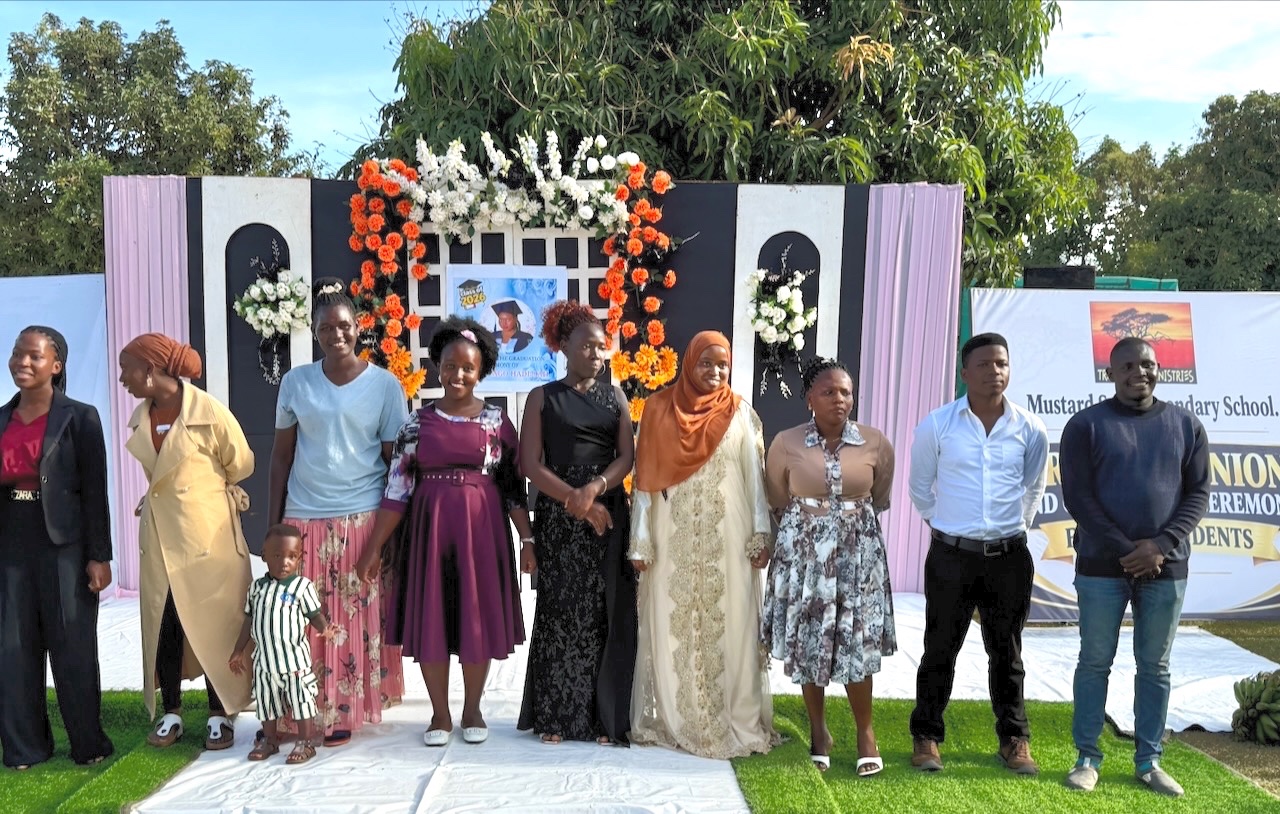Nine smiling young adults and one small child lined up in front of a colorful background.