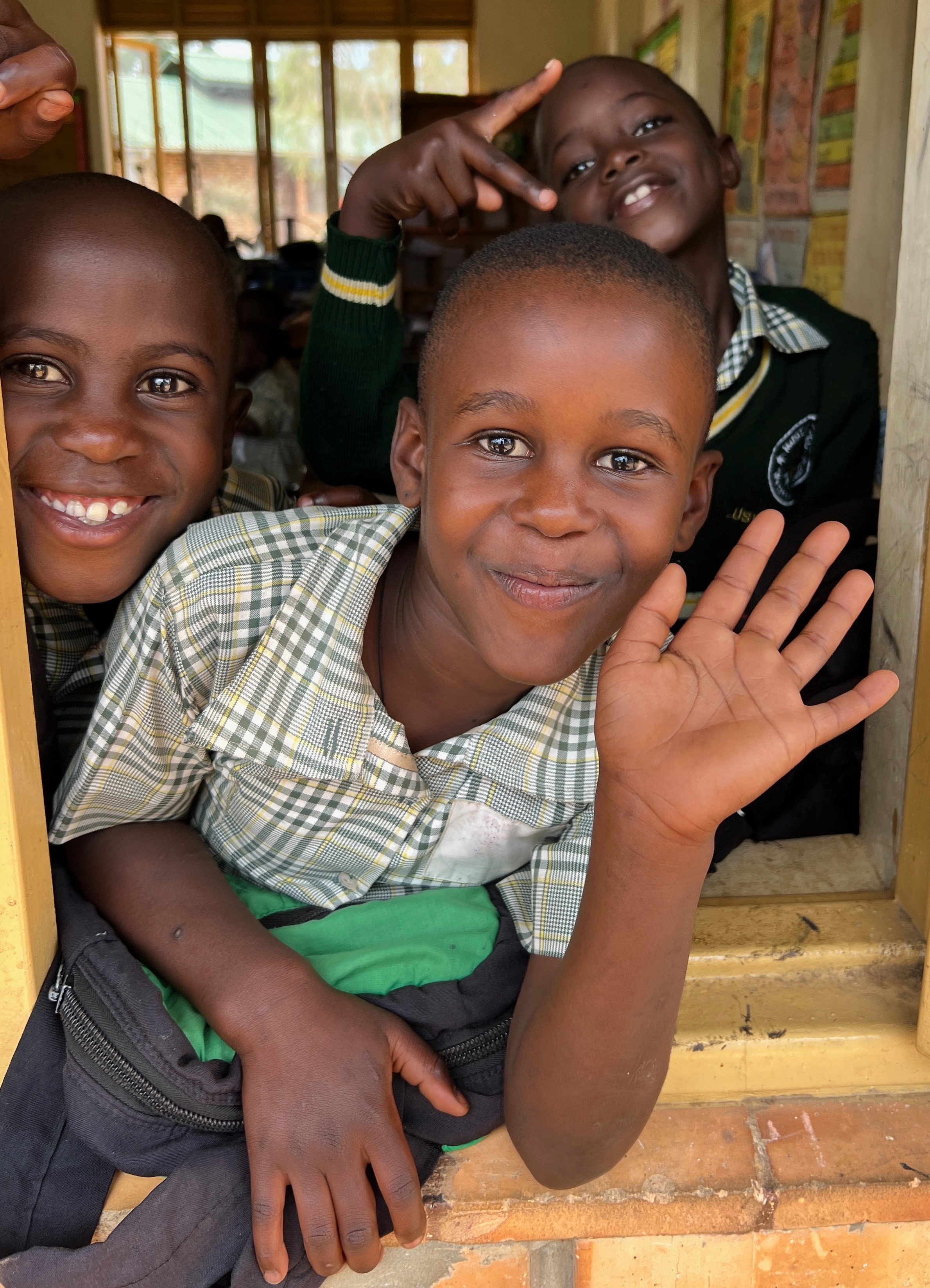 Three third-grade Ugandan boys in school uniforms are greeting visitors through an open lassroom window