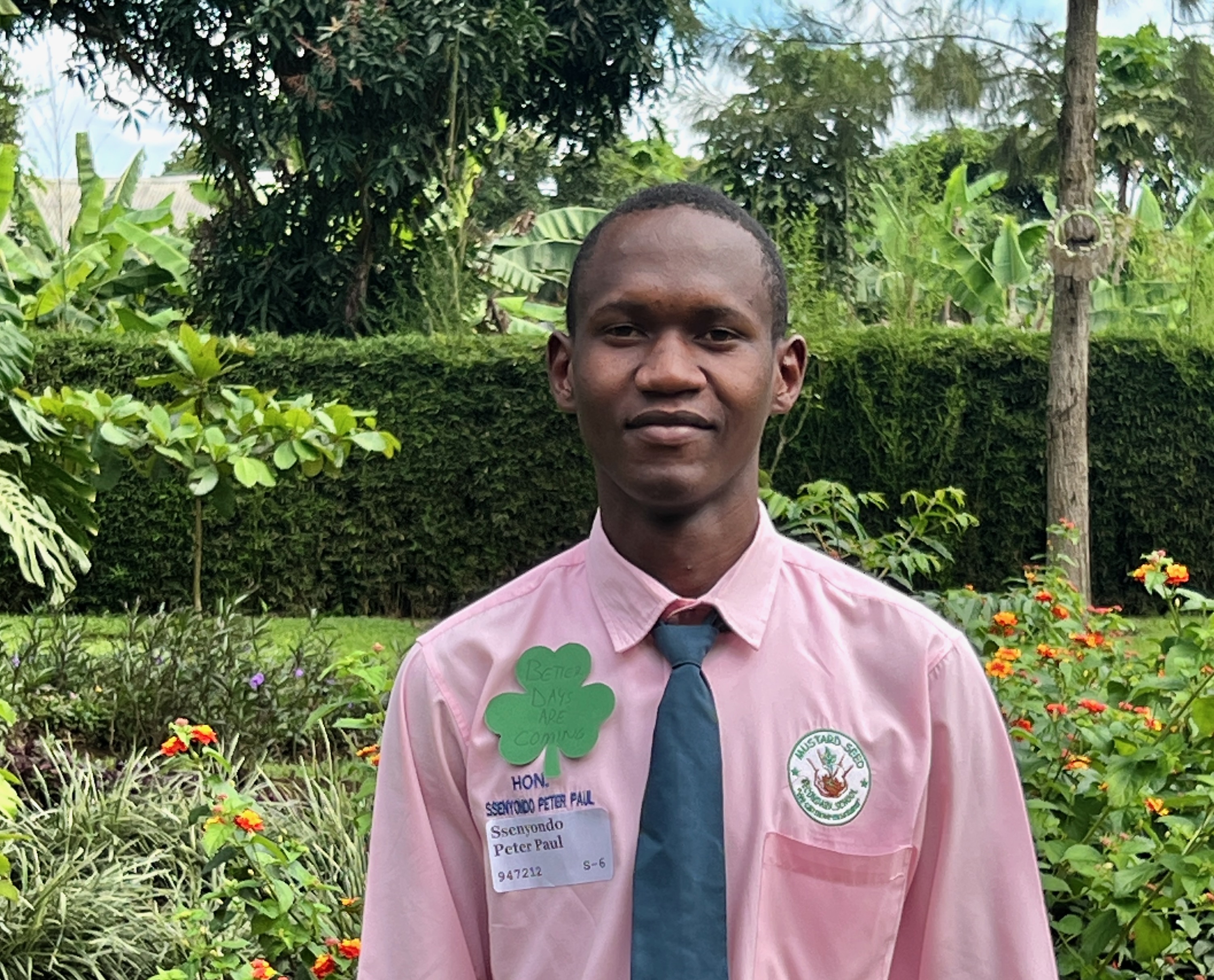 A twenty year old Ugandan student, dressed in a pink dress shirt with a teal green tie, with green campus in the background
