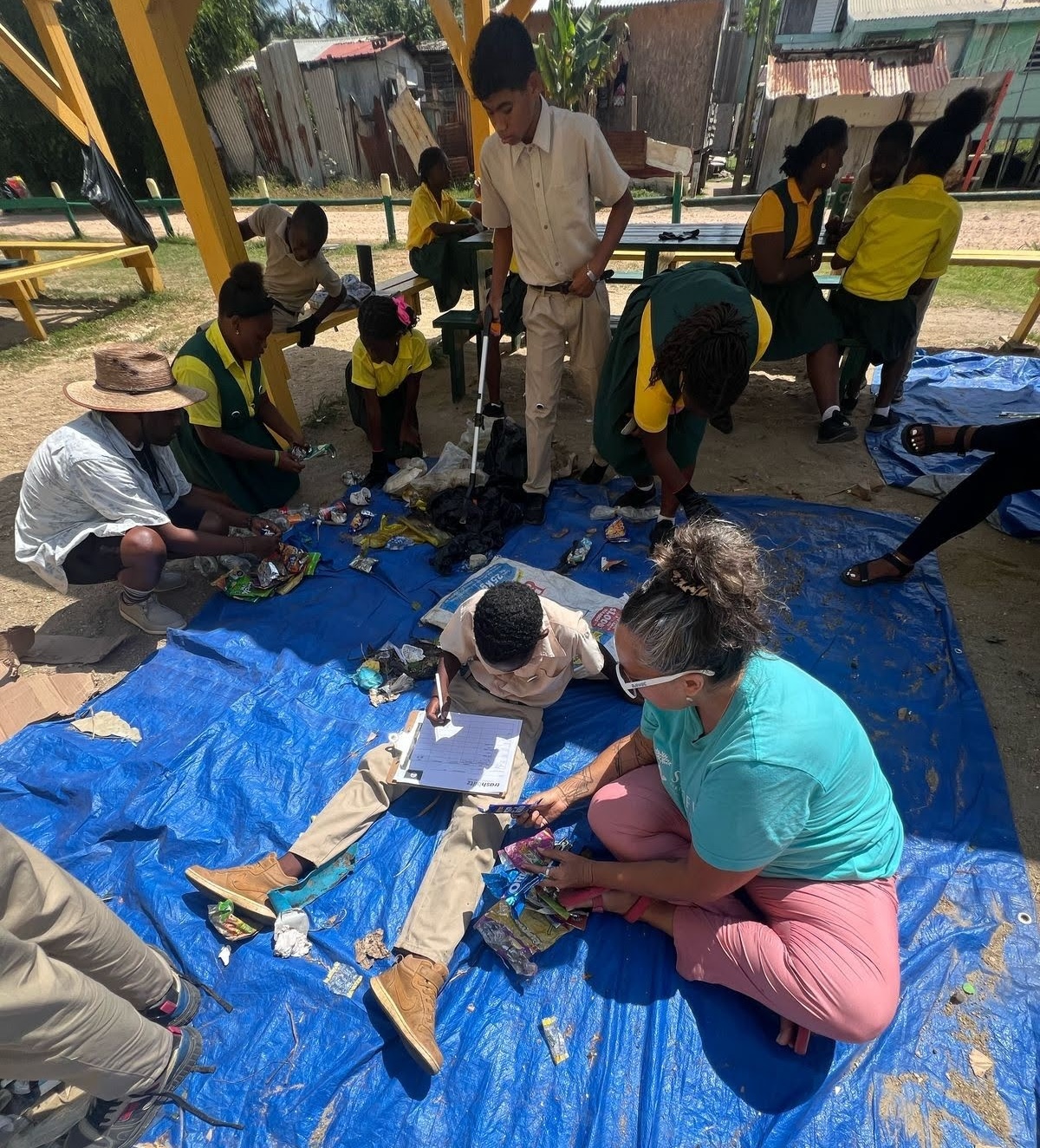 A group of children and adults sit and stand on a blue tarp outdoors, sorting plastic materials. 