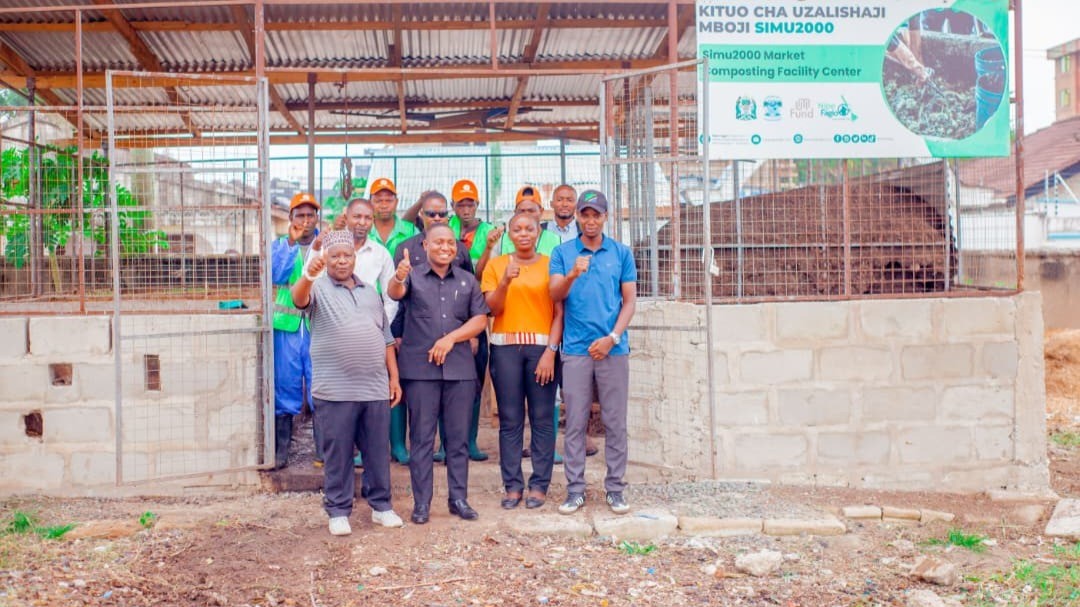 A group of people stands smiling in front of a composting facility, giving thumbs up. 