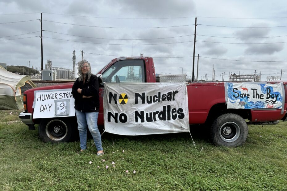 Diane Wilson stands by a red truck displaying banners reading “Hunger Strike Day 9,“ “No Nuclear No Nurdles,“ and “Save The Bay“ on a grassy area near industrial structures.