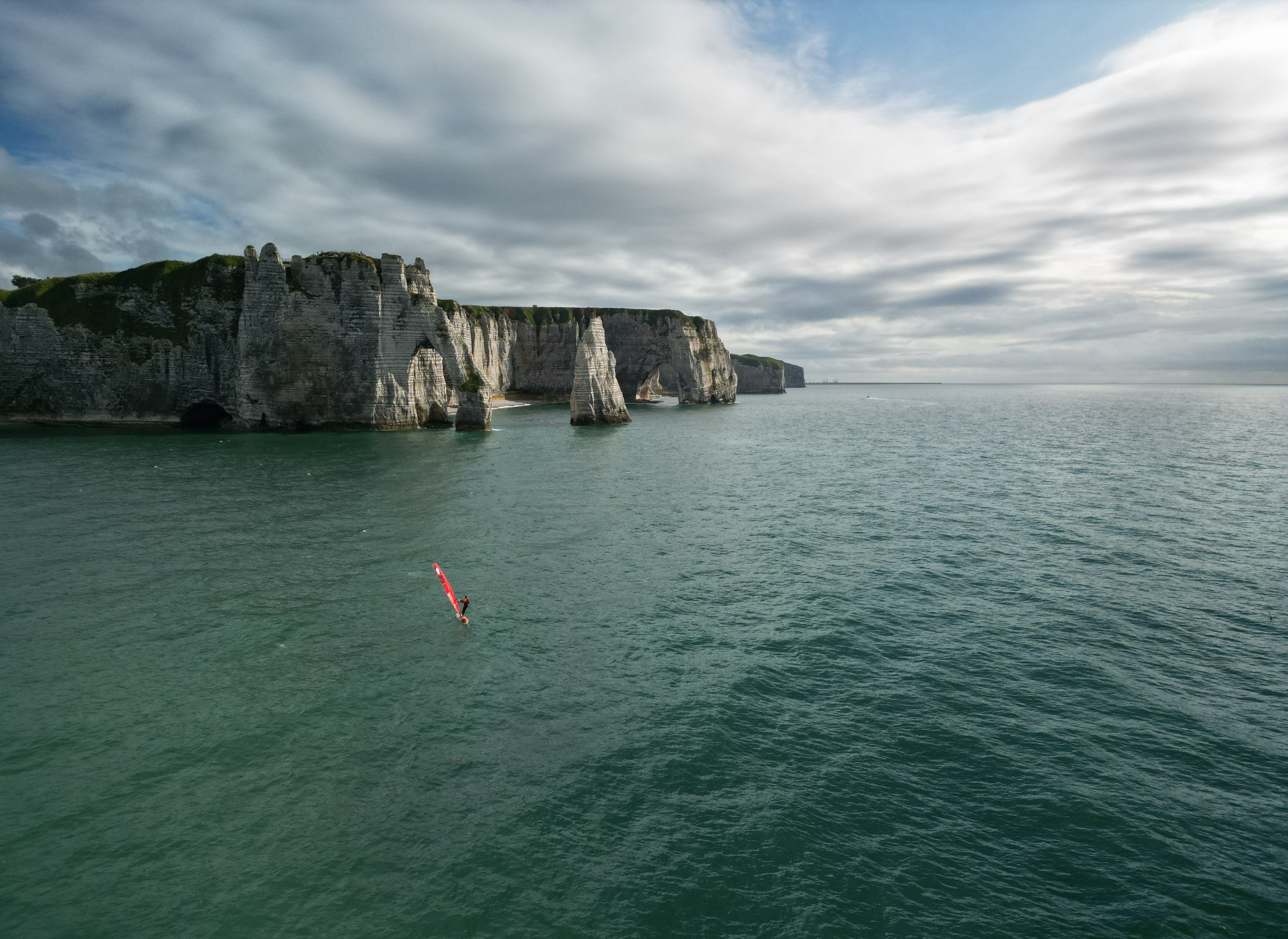 A lone windsurfer glides on calm turquoise waters near towering white limestone cliffs under a sky with scattered clouds.