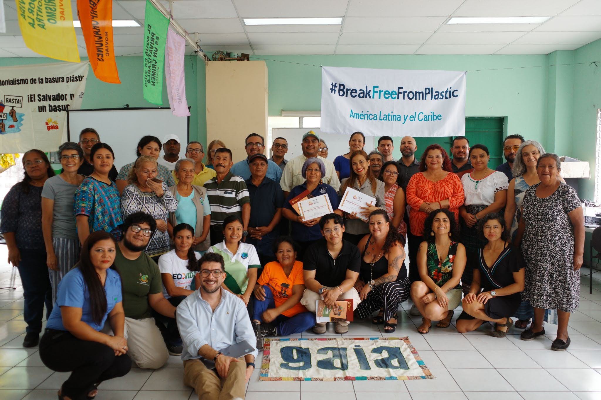 A diverse group of about 35 people pose indoors, smiling, with a banner reading “#BreakFreeFromPlastic América Latina y el Caribe“ above. 