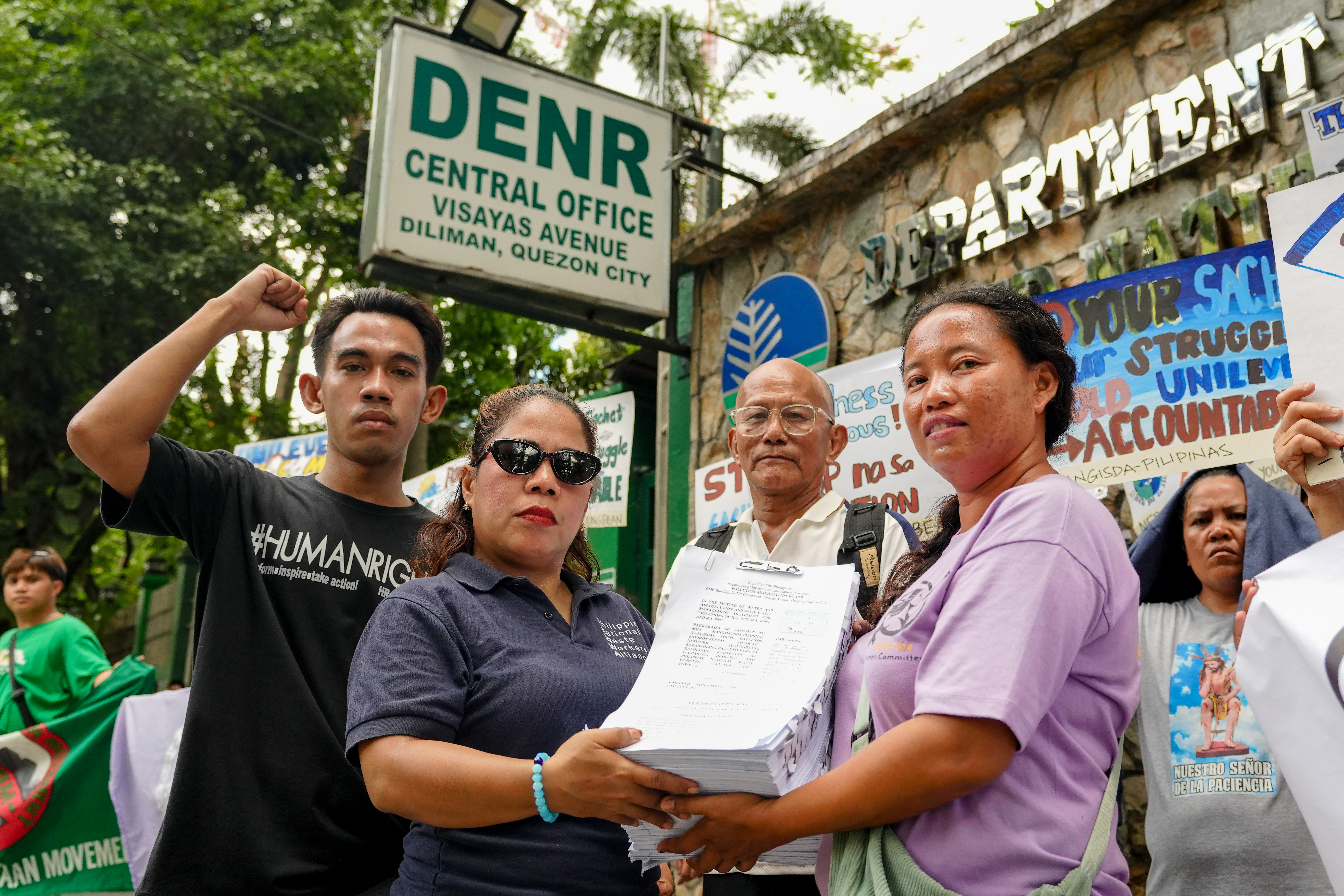 A group of protesters stands outside the DENR Central Office, holding a stack of papers and signs. 
