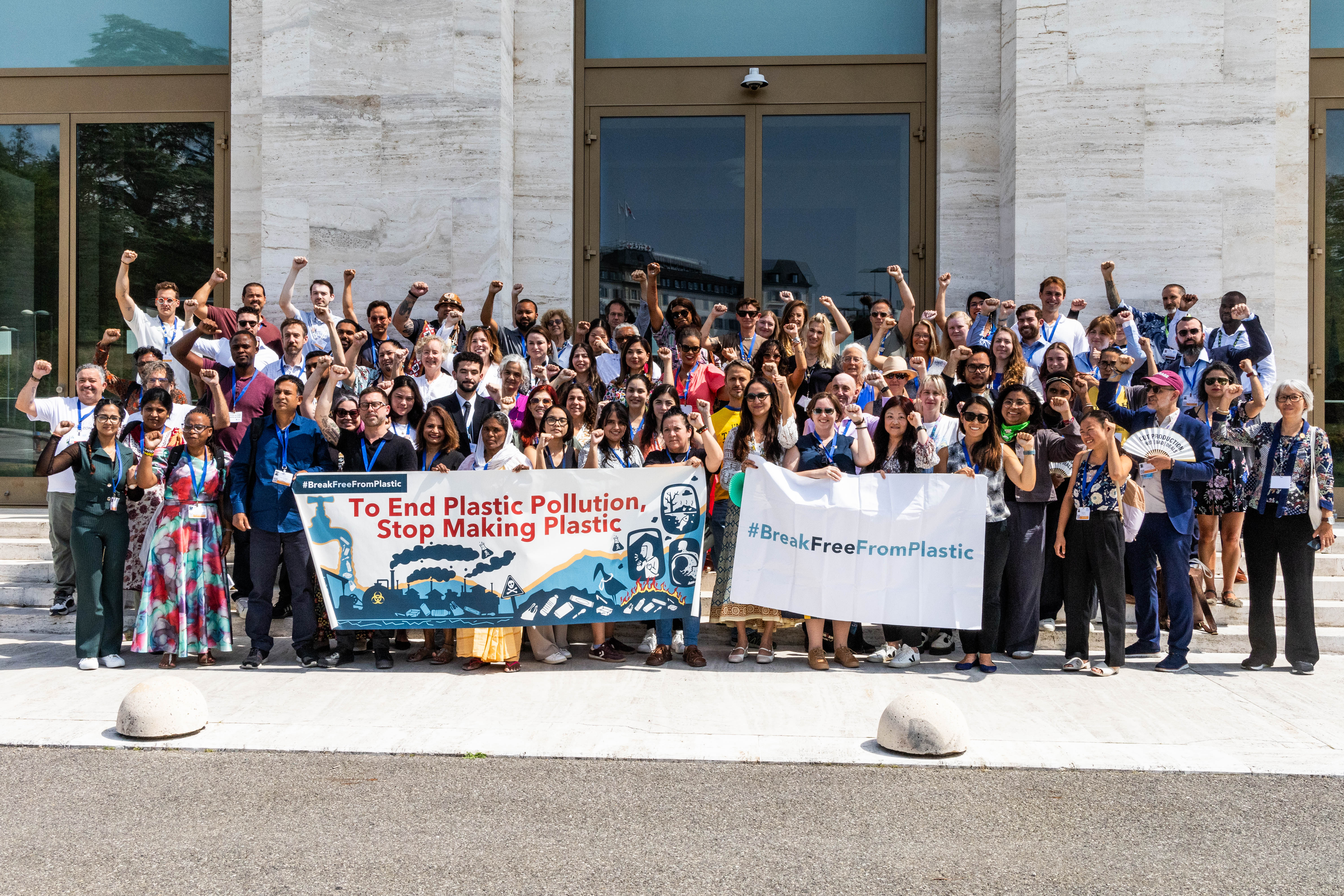 A large, diverse group of people hold signs reading “To End Plastic Pollution, Stop Making Plastic“ and “#BreakFreeFromPlastic“ outside a modern building.
