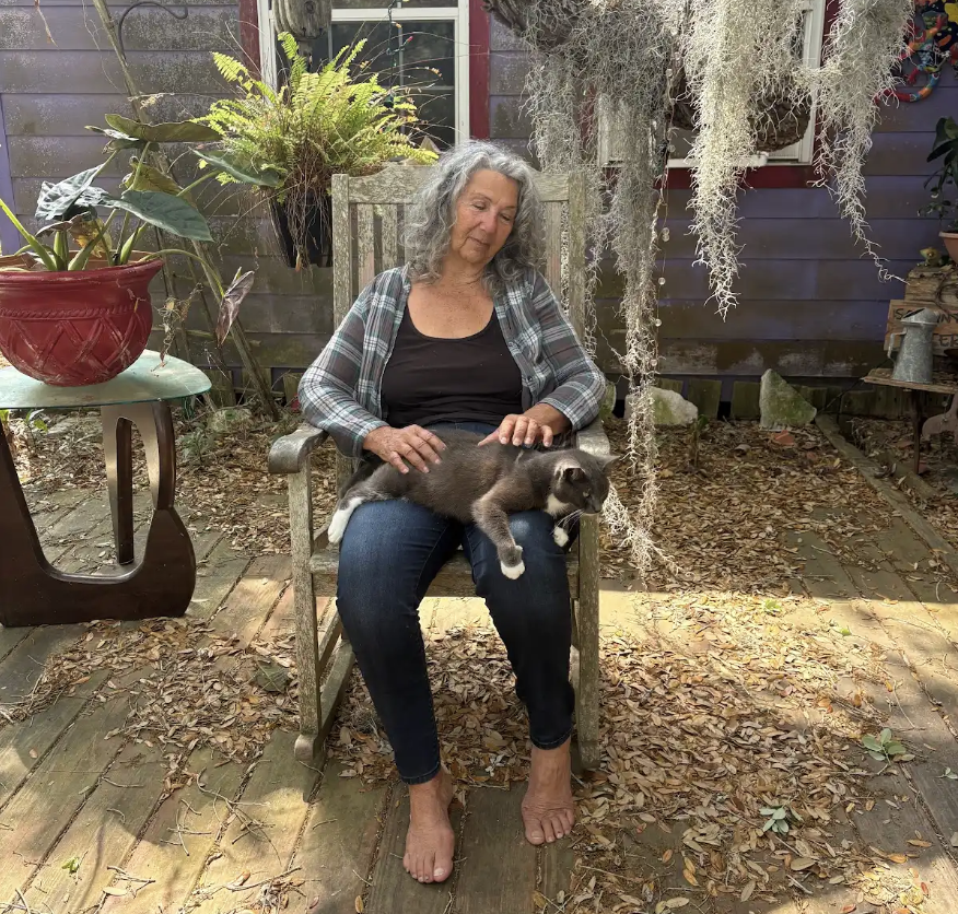 A woman with long gray hair sits on a wooden chair on a leafy porch, gently petting a relaxed cat on her lap. 