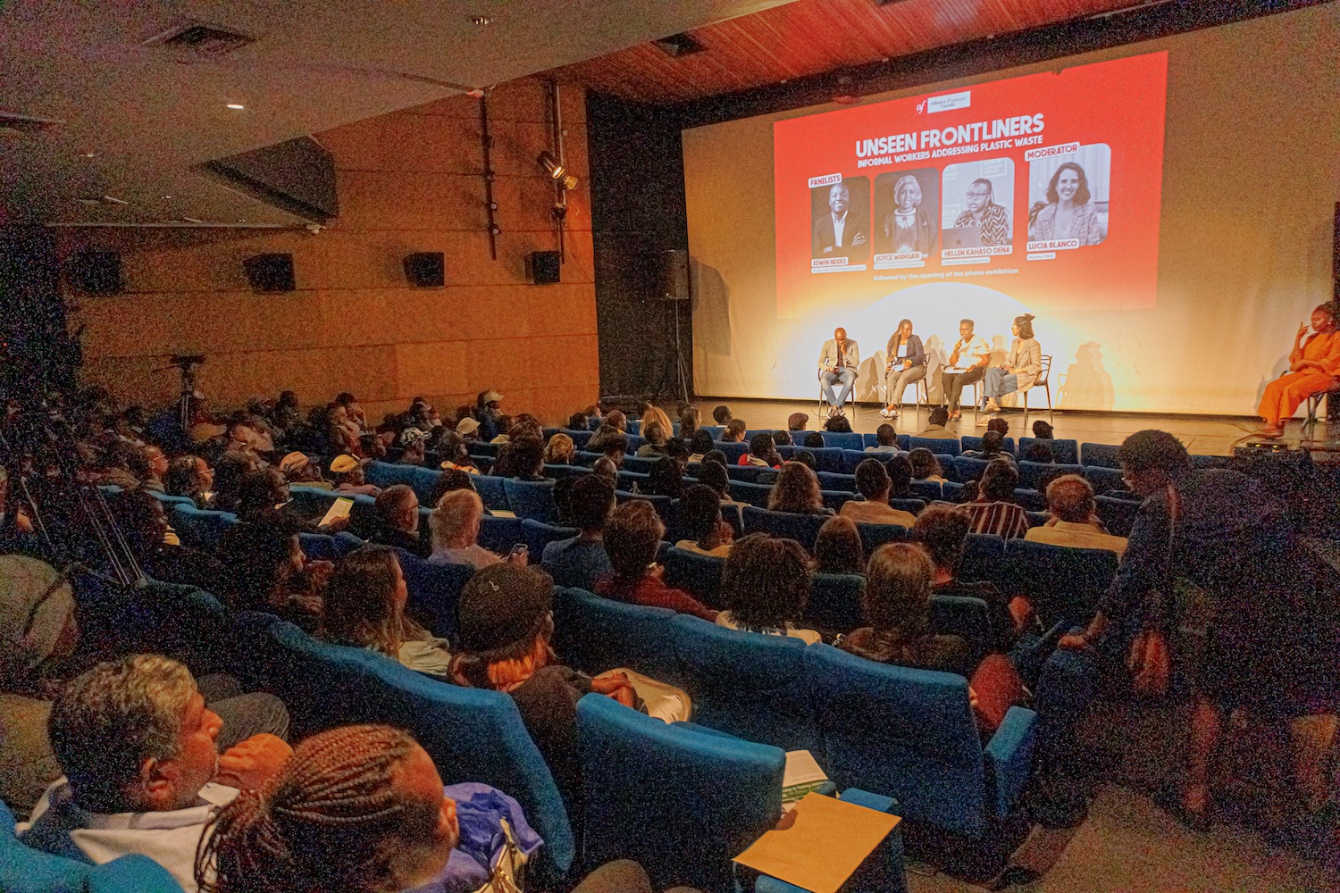 Audience seated in a theater with blue chairs, watching a panel discussion titled “Unseen Frontliners.“ Four panelists are on stage, engaged in conversation. 